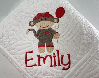Puede incluir: Manta de bebé acolchada blanca con un mono bordado en marrón y rojo que sostiene un globo rojo. El nombre "Emily" está bordado en rojo debajo del mono.