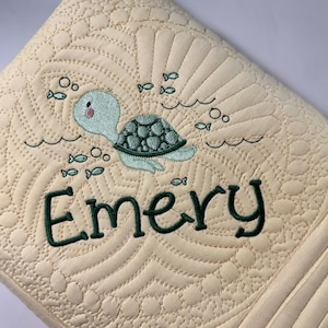 May include: Cream-colored, quilted blanket with an embroidered sea turtle design. The turtle is light blue and green, swimming with fish and bubbles. The name "Emery" is embroidered in dark green script below the turtle.