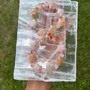 May include: A gemstone bracelet featuring star-shaped and round beads in shades of peach, pink, and orange. The bracelet is displayed on a clear, rectangular crystal. The background is green grass.