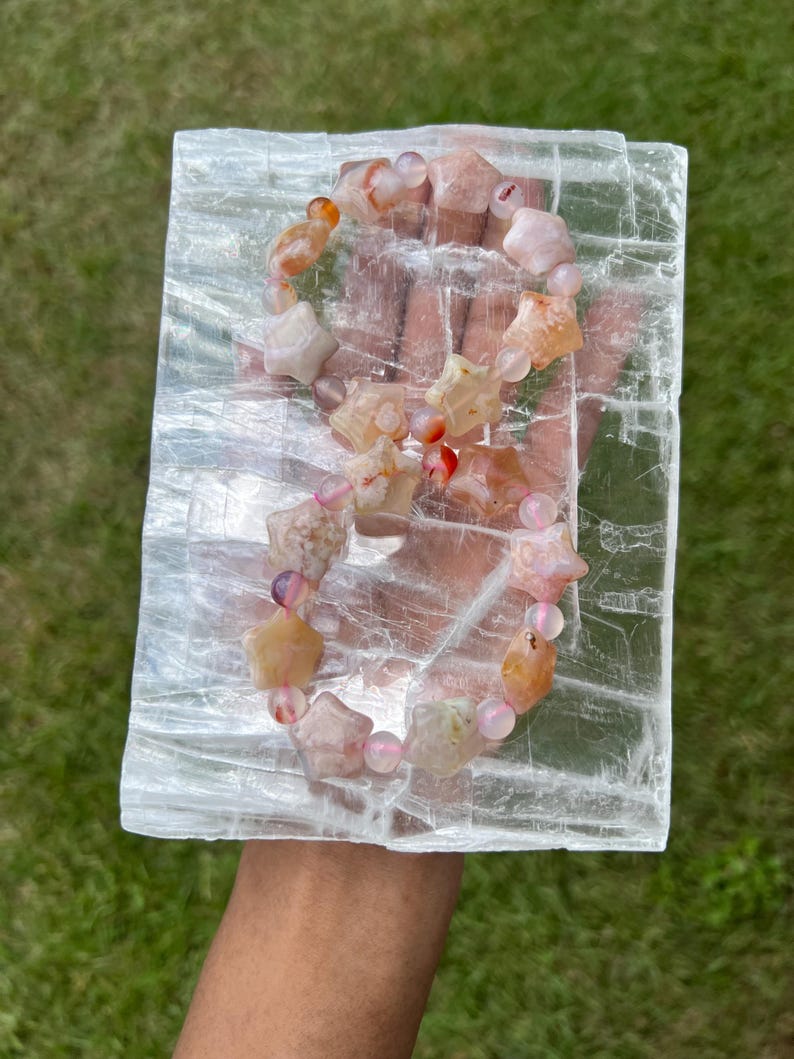 May include: A gemstone bracelet featuring star-shaped and round beads in shades of peach, pink, and orange. The bracelet is displayed on a clear, rectangular crystal. The background is green grass.