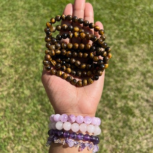 May include: A hand displays several beaded bracelets. The top bracelets are brown and gold tiger's eye. Below are bracelets in shades of lavender and purple, including amethyst and clear crystal accents. The background is green grass.