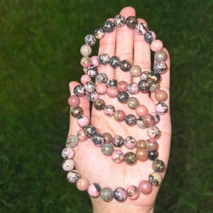 May include: A beaded necklace featuring a variety of round, polished stones in shades of pink, black, and brown. The necklace is held in a hand against a green background. The beads appear to be approximately 0.3 inches in diameter.