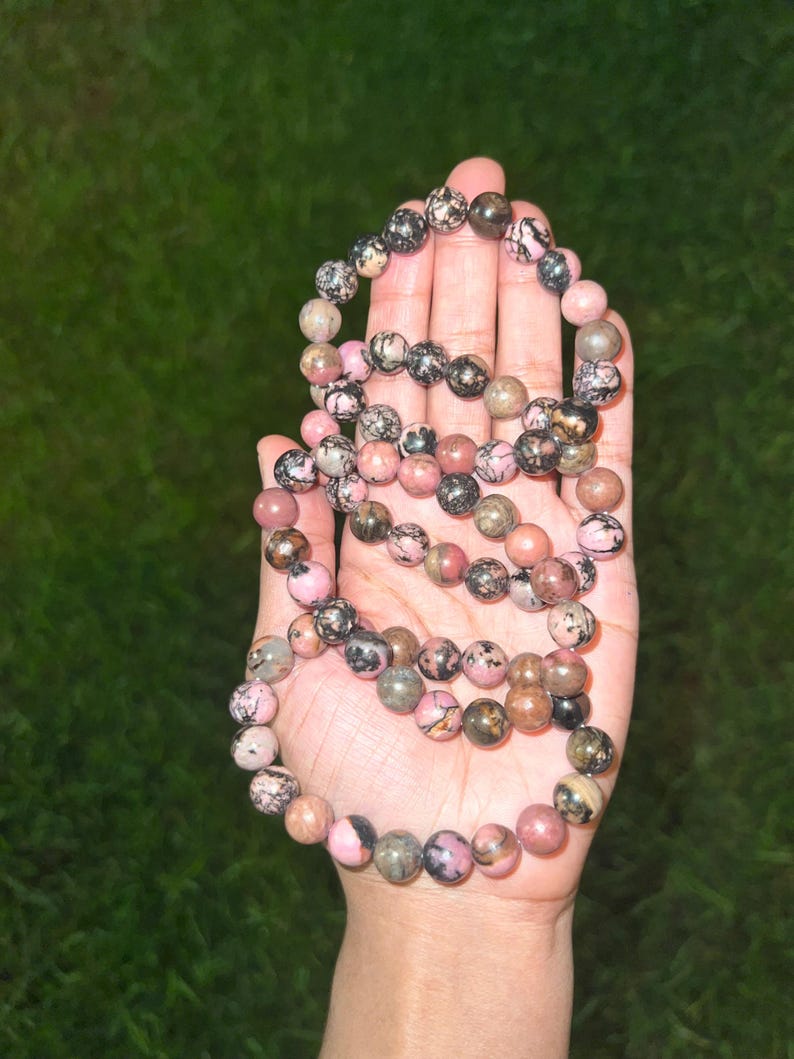 May include: A beaded necklace featuring a variety of round, polished stones in shades of pink, black, and brown. The necklace is held in a hand against a green background. The beads appear to be approximately 0.3 inches in diameter.
