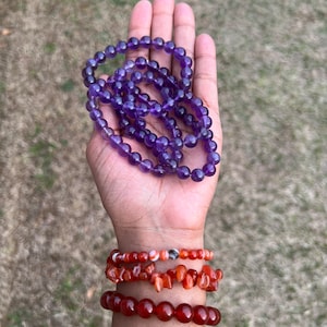 May include: Two bracelets made of amethyst and carnelian beads. The amethyst bracelet is purple and the carnelian bracelet is orange. The bracelets are stacked on a person's wrist.