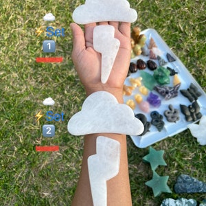 May include: Two sets of white cloud and lightning shaped crystal gua sha tools. Set 1 and Set 2 are labeled on the image. The tools are on a hand and a white tray with other crystals.