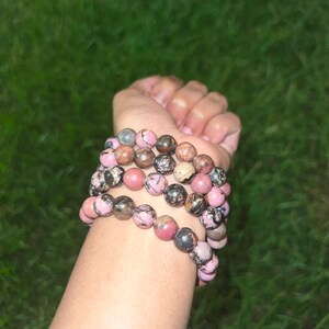 May include: Several beaded bracelets with pink, brown, and gray stones. The bracelets are stacked on top of each other. The beads are round and appear to be made of natural stone.
