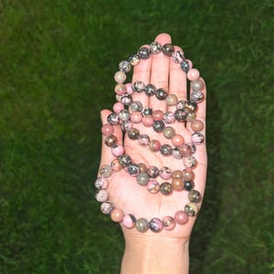 May include: A beaded necklace with pink, black, and gray stones. The necklace is held in an open hand against a green background. The beads are round and vary in size, with a mix of colors and patterns.