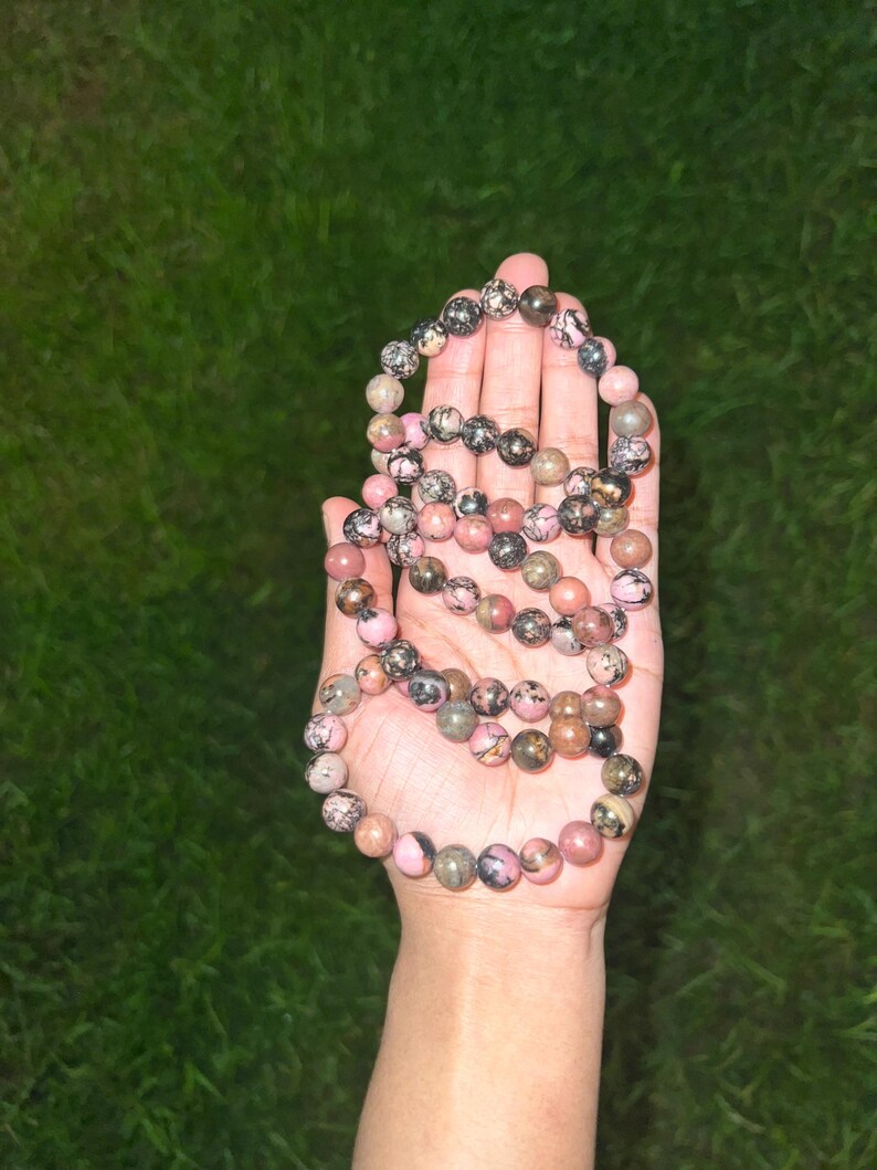 May include: A beaded necklace with pink, black, and gray stones. The necklace is held in an open hand against a green background. The beads are round and vary in size, with a mix of colors and patterns.