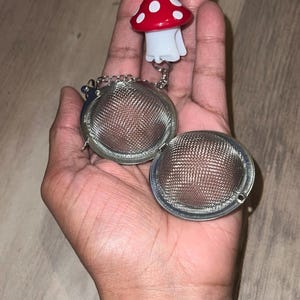May include: A tea infuser with a mushroom-shaped top. The mushroom has a red cap with white spots and a white stem. The infuser is made of a fine mesh and opens in two halves, connected by a chain.