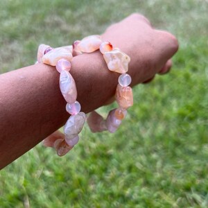 May include: A bracelet featuring a mix of peach and pink-colored, irregularly shaped gemstone beads. The bracelet is strung together with smaller, round, pink beads. The gemstones have a natural, organic appearance, with variations in color and texture.