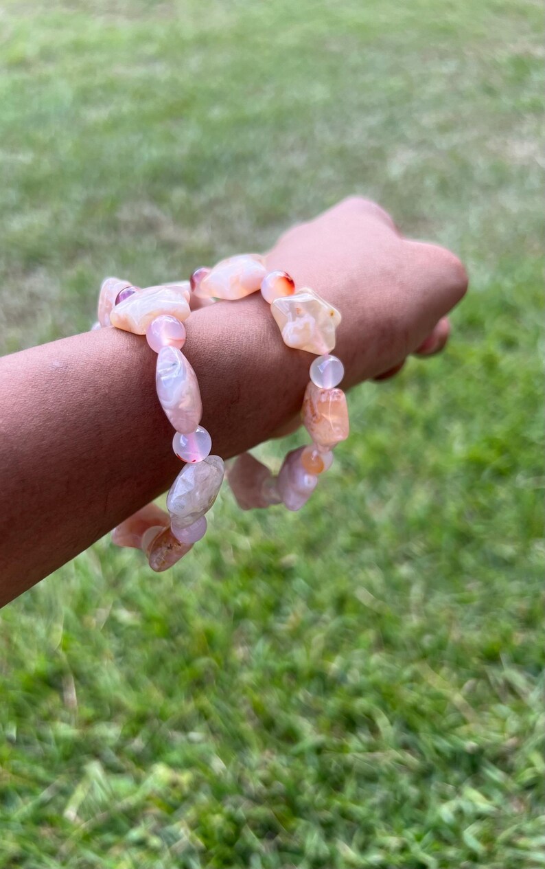 May include: A bracelet featuring a mix of peach and pink-colored, irregularly shaped gemstone beads. The bracelet is strung together with smaller, round, pink beads. The gemstones have a natural, organic appearance, with variations in color and texture.