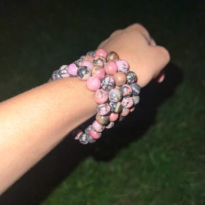 May include: A collection of beaded bracelets in shades of pink, brown, and gray. The bracelets are made of round, polished stones and are stacked on a wrist. The background is dark, with a hint of green grass.
