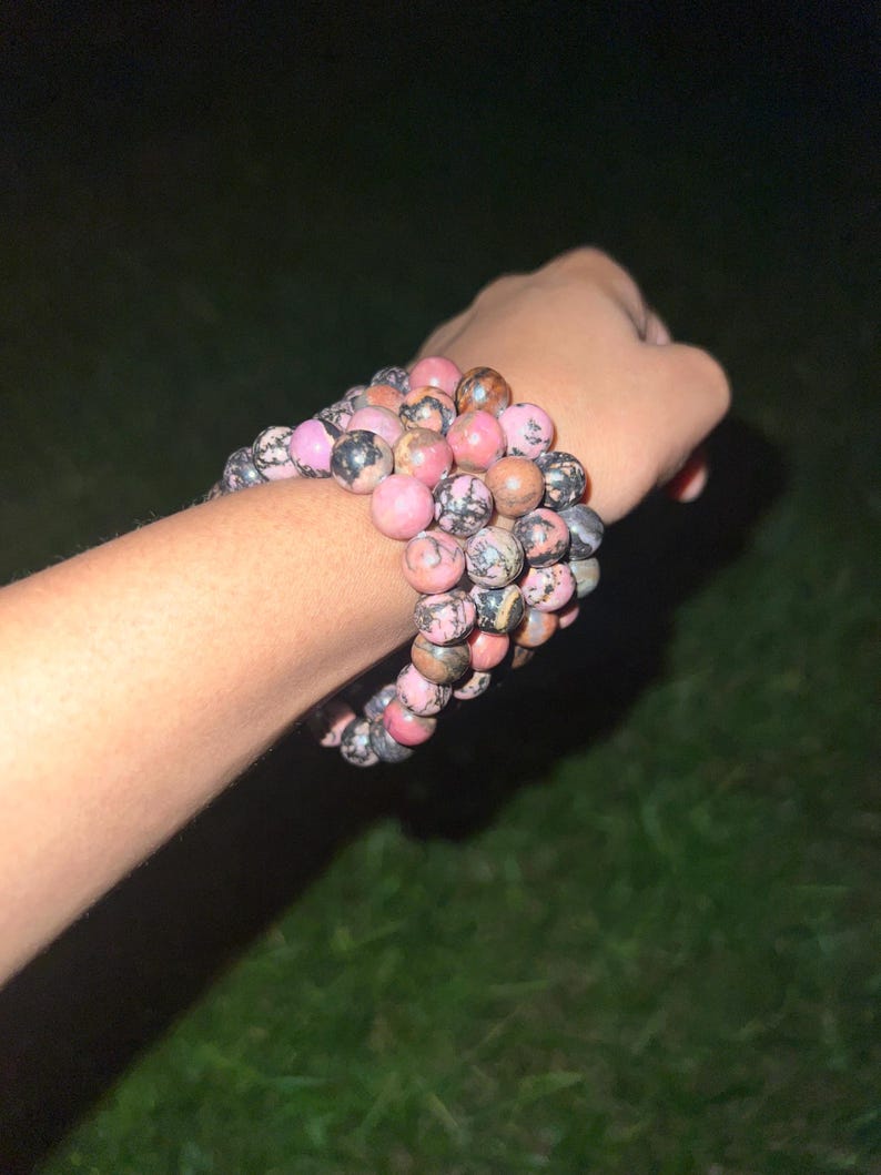 May include: A collection of beaded bracelets in shades of pink, brown, and gray. The bracelets are made of round, polished stones and are stacked on a wrist. The background is dark, with a hint of green grass.