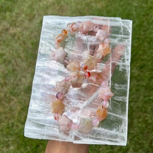 May include: A gemstone bracelet with star-shaped beads in shades of peach, pink, and orange, interspersed with round pink beads. The bracelet rests on a rectangular, translucent white crystal against a backdrop of green grass.