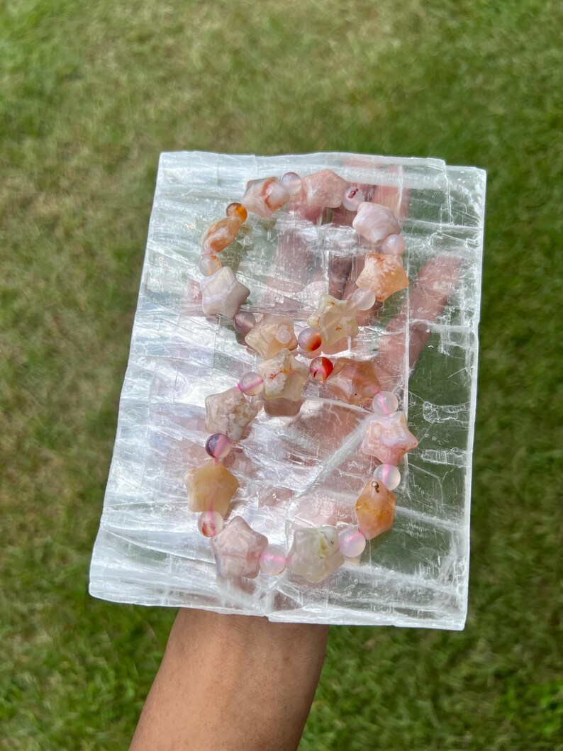 May include: A gemstone bracelet with star-shaped beads in shades of peach, pink, and orange, interspersed with round pink beads. The bracelet rests on a rectangular, translucent white crystal against a backdrop of green grass.