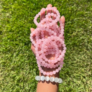 May include: A stack of pink rose quartz bracelets with a white moonstone bracelet on top. The bracelets are made of round beads and are displayed on a hand against a green grass background.