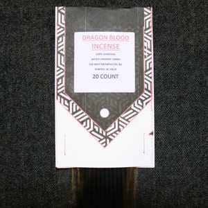 May include: A white package with black and white geometric designs. The package contains 20 Dragon Blood incense sticks. The package label reads "Dragon Blood Incense", "100% Charcoal", "Asya's Organic Farms", "320 West Brewington Rd.", "Sumter, SC 29153", and "20 Count".