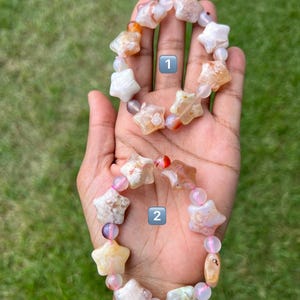 May include: Two star-shaped gemstone bracelets. One bracelet features white, orange, and tan star beads with pink and purple accent beads. The other bracelet has similar star beads with pink and purple accent beads. Both bracelets are on a hand.