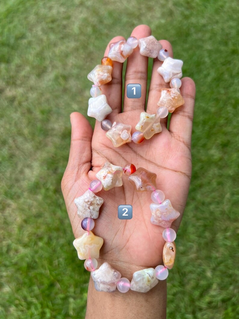 May include: Two star-shaped gemstone bracelets. One bracelet features white, orange, and tan star beads with pink and purple accent beads. The other bracelet has similar star beads with pink and purple accent beads. Both bracelets are on a hand.