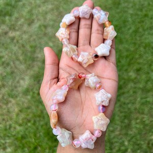 May include: Two star-shaped bracelets with a mix of white, orange, and pink beads. The bracelets are held in a hand against a backdrop of green grass. The star beads are the focal point of the bracelets.