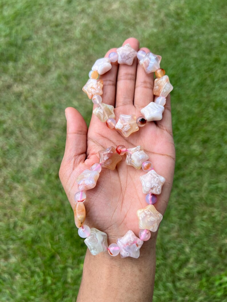 May include: Two star-shaped bracelets with a mix of white, orange, and pink beads. The bracelets are held in a hand against a backdrop of green grass. The star beads are the focal point of the bracelets.