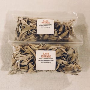 Può includere: Due sacchetti di plastica trasparente pieni di foglie di salvia bianca essiccate. I sacchetti hanno etichette che recitano "Sage Incense" e includono il nome dell'azienda, l'indirizzo e le informazioni di contatto.