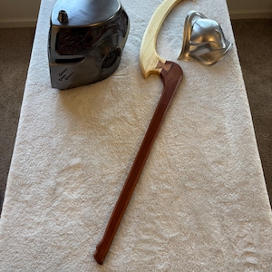 May include: A dark metal helmet and a wooden and metal weapon are displayed on a white surface. The helmet has intricate details and a small nozzle on top. The weapon features a wooden handle and a curved blade.