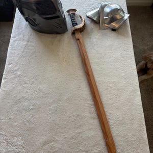 Puede incluir: Un conjunto de objetos de temática medieval, que incluye un casco plateado, una espada de madera con empuñadura curva y una pieza de armadura de hombro plateada. La espada es de madera marrón claro y tiene una empuñadura negra.