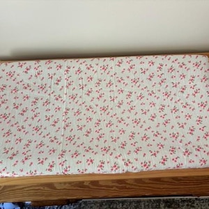 May include: A rectangular baby changing pad with a white background and a pattern of small pink flowers. The pad is set inside a wooden frame, suggesting a changing table or similar furniture.