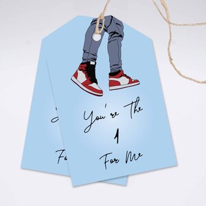 May include: Two pale blue gift tags featuring an illustration of legs in grey jeans and red, white, and black trainers. The tags have the text "You're the 1 For Me" in black script.