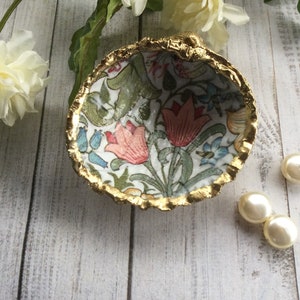 May include: A small, white ceramic dish with a gold rim and a floral pattern. The dish is filled with three white pearl-like beads.