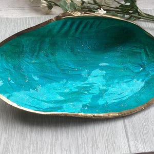 May include: A teal blue seashell dish with a gold rim. The dish is made of resin and has a sparkly finish.