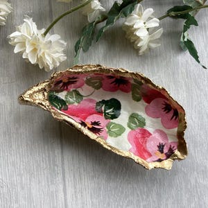 May include: A decorative oyster shell dish with a gold-painted rim. The interior features a floral design with pink and red pansies and green leaves on a white background. The shell is placed on a light gray wooden surface.