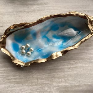 May include: An oyster shell trinket dish with a gold-painted edge. The interior has a blue and white marbled effect, with a small white seashell and a cluster of pearls. A unique coastal-themed decorative item.