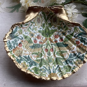 May include: A ceramic dish shaped like a seashell with a gold rim, featuring a white background and a floral pattern with birds, leaves, and strawberries.