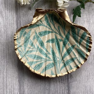 May include: A decorative seashell trinket dish. The interior has a cream background with a teal leaf pattern. The rim is trimmed with gold paint. The dish is on a grey wooden surface.