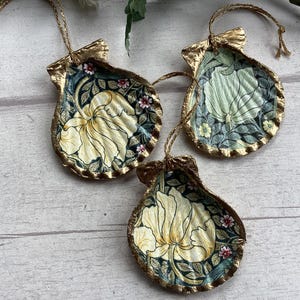 May include: Three seashell ornaments with gold trim and floral designs. Each shell features a different floral pattern in shades of cream, green, and pink against a dark blue background. The ornaments are suspended by gold cords.