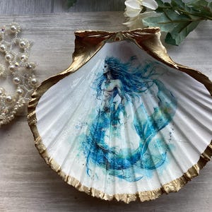 May include: A large, white seashell with a gold-painted rim and a watercolor painting of a mermaid inside. The mermaid has long blue hair and a blue tail. A pearl necklace is next to the shell.