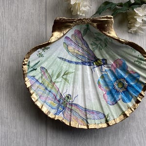 May include: A decorative scallop shell dish featuring a light green background with two colourful dragonflies and a large blue flower. The edges of the shell are painted with a metallic gold finish, creating a distinct border around the interior design.