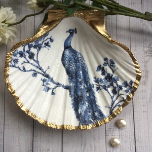 May include: A decorative seashell dish with a gold-painted rim. The interior features a blue and white peacock design. Two small, round, white pearls are next to the dish. The dish is on a light wood surface.