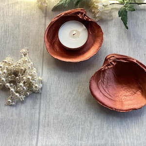 May include: Two copper-coloured painted seashells, one containing a white tea light candle, and a cluster of small white faux pearl beads. The shells have a metallic finish and a smooth, concave interior surface for holding small items or candles.