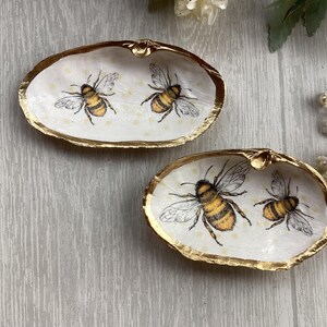 May include: Two white seashells with gold trim, each featuring a painted design of bees. The bees are yellow and black with brown wings.