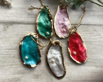 May include: Six decorative oyster shell ornaments with gold trim and a pearl accent. The shells are painted in various colors, including teal, green, pink, red, and white. Each shell has a gold string for hanging.