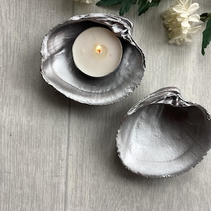 May include: Two natural clam shells painted with a metallic silver finish. One shell holds a small, round white tea light candle with a lit flame. The interior of both shells is smooth and reflective, showcasing the silver paint application.