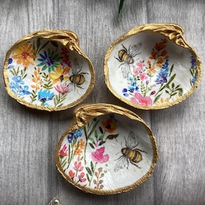 May include: Three decorative seashell dishes with gold-painted edges. Each shell is hand-painted with colorful flowers and a bee. The shells are arranged on a gray wooden surface, showcasing a nature-inspired theme.