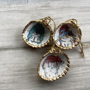 May include: Three seashell ornaments with gold-painted edges and gold string hangers. Each shell features a hand-painted cat wearing a winter hat. The hats are teal, red, and blue. These ornaments are perfect for cat lovers.