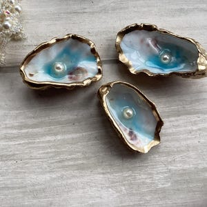 May include: Three decorative oyster shell trinket dishes. Each shell has a blue and white painted interior and a gold-coloured rim. A single pearl sits in the centre of each shell. A beaded pearl strand is visible in the upper left corner.