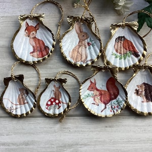 May include: Set of eight white seashell ornaments with gold trim. Each ornament features a different woodland animal illustration, including a fox, rabbit, owl, hedgehog, squirrel, and two mice.