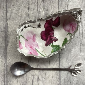 May include: A silver bowl with a floral design and the text "Save the bees..."  A silver spoon with a leaf design is next to the bowl.