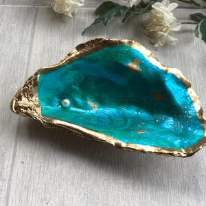 May include: An oyster shell dish with a vibrant turquoise interior featuring gold leaf accents and a single small white pearl resting inside. The outer edge of the shell is painted with a metallic gold finish, creating a decorative bowl.